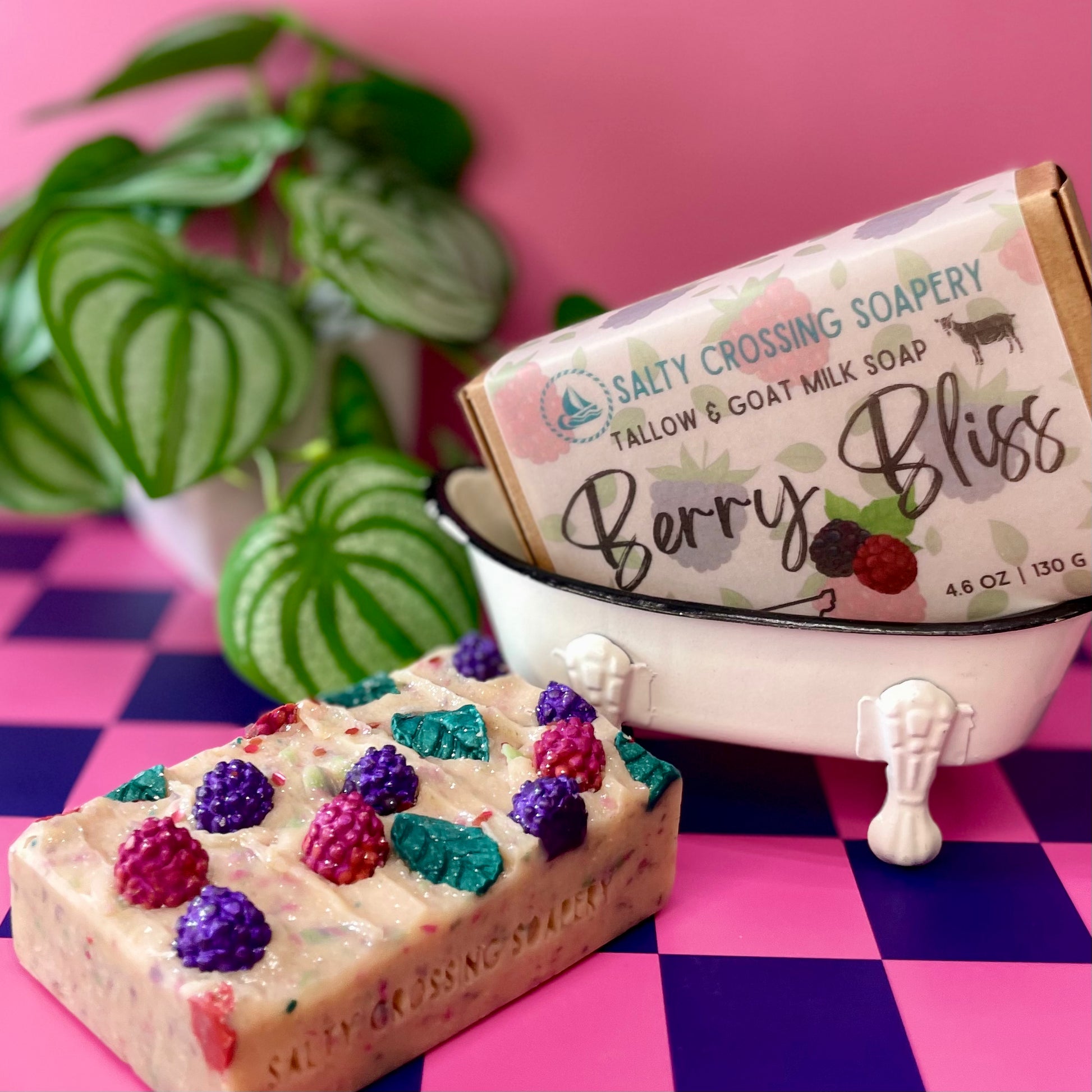 Bar of soap with berry decorations in front of a miniature bathtub and 'Salty Crossing Soapery' packaging on a checkered background.