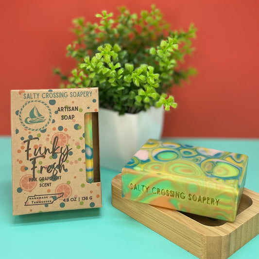 Salty Crossing Soapery soap bar and packaging on a teal surface with a plant in the background.