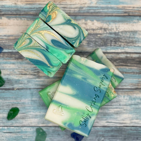 Arrangement of soap bars. They are swirled with green and white, with a touch of gold on the top. The bar face is stamped with a small sailboat graphic and the text salty crossing soapery