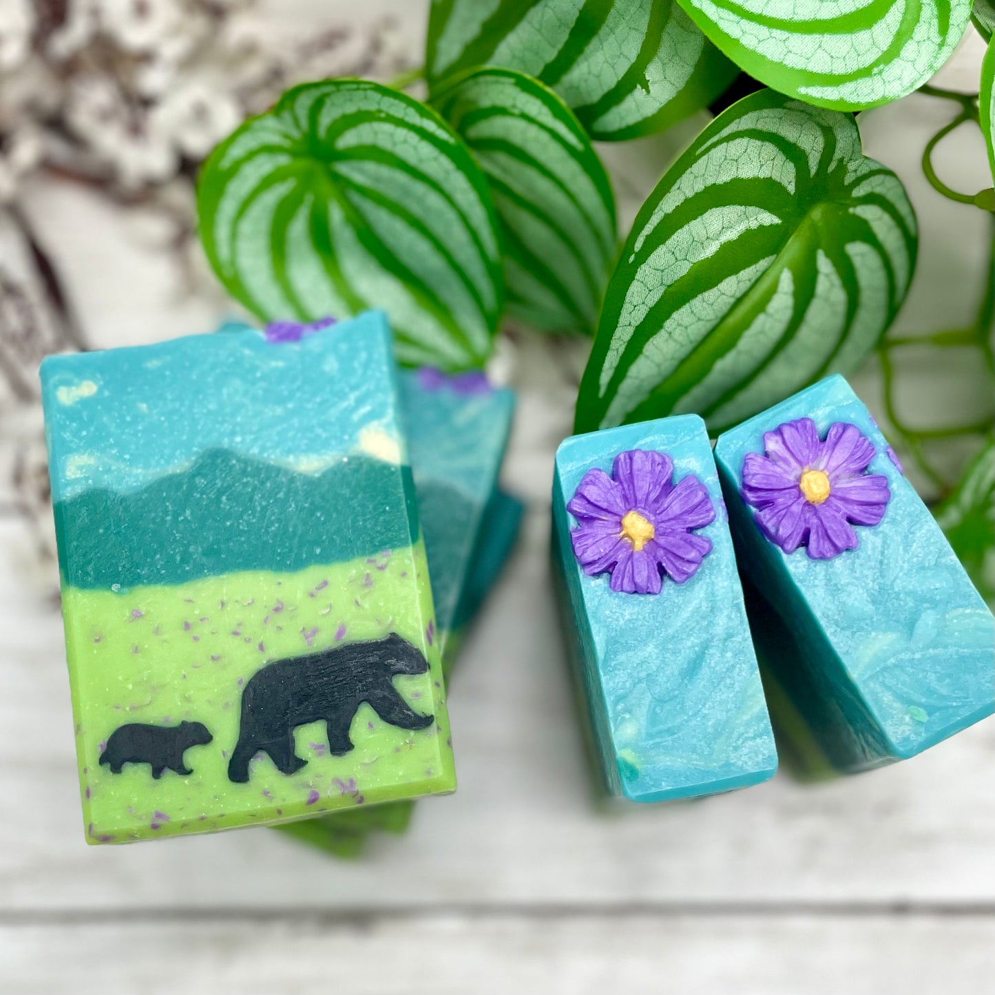Three soap bars with nature-themed designs on a wooden surface with green leaves in the background.