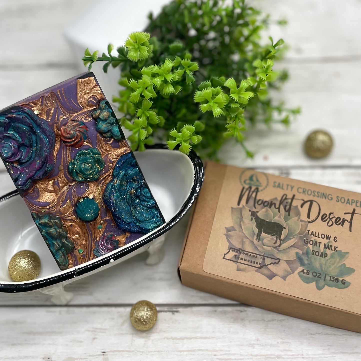 Decorative soap bar with floral design in a black dish, next to a box of 'Moonlit Desert' soap on a wooden surface.