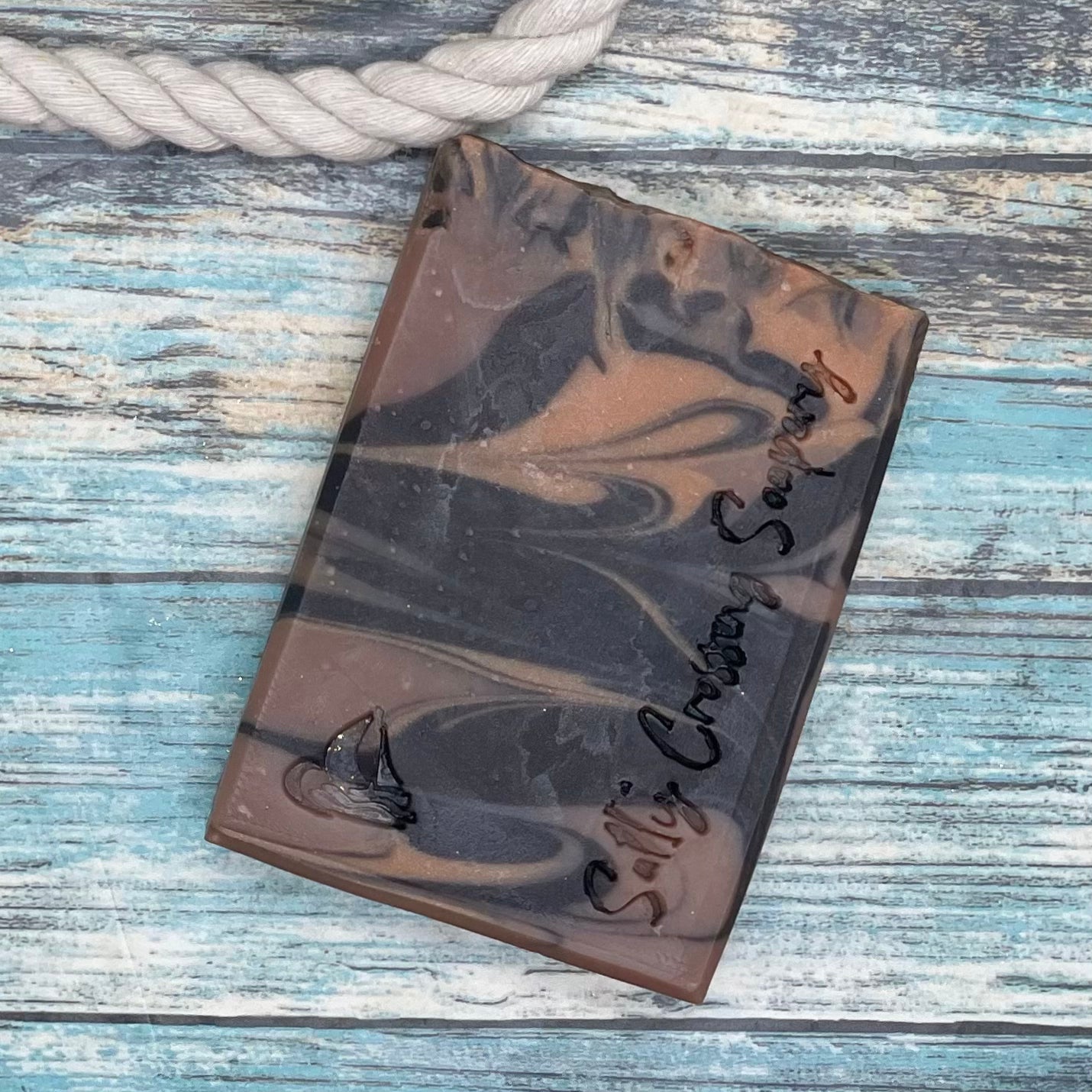 Smokin jack soap for men. Close up of a bar which is swirled in 3 shades of brown, then handstamped with a small sailboat graphic and the text Salty Crossing Soapery