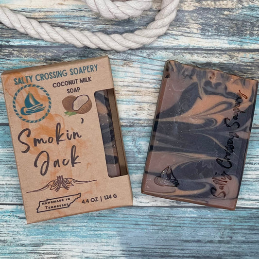 Smokin jack soap for men. Bar of soap with its box. The soap is dark chocolate brown, medium brown, and caramel colored swirls. It’s stamped with a small sailboat graphic and the text Salty Crossing Soapery. The box is kraft paperboard printed with color text and graphics.