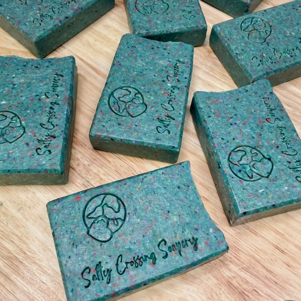 Multiple bars of blue soap with speckles on a wooden surface, branded 'Sally Crossing Soapery'.
