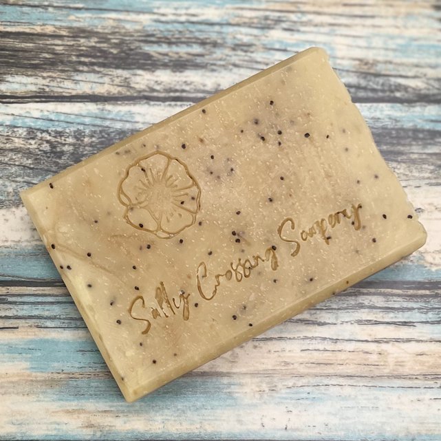 close up detail of soap bar. light yellow color with poppy seeds throughout. stamped with a poppy flower and Salty Crossing Soapery.